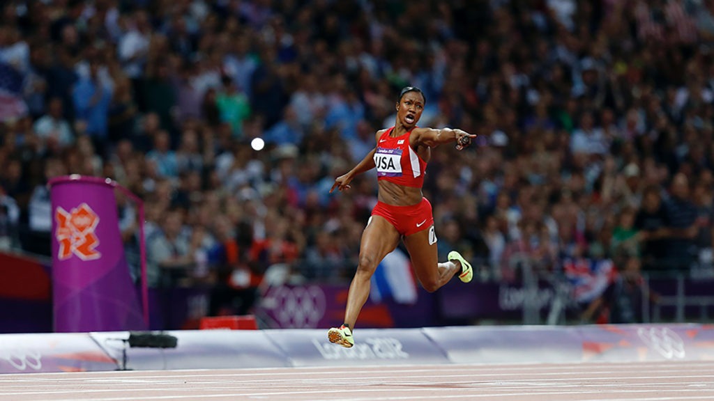Carmelita Jeter extends her left arm hoding the baton and points at the world-record time on a scoreboard